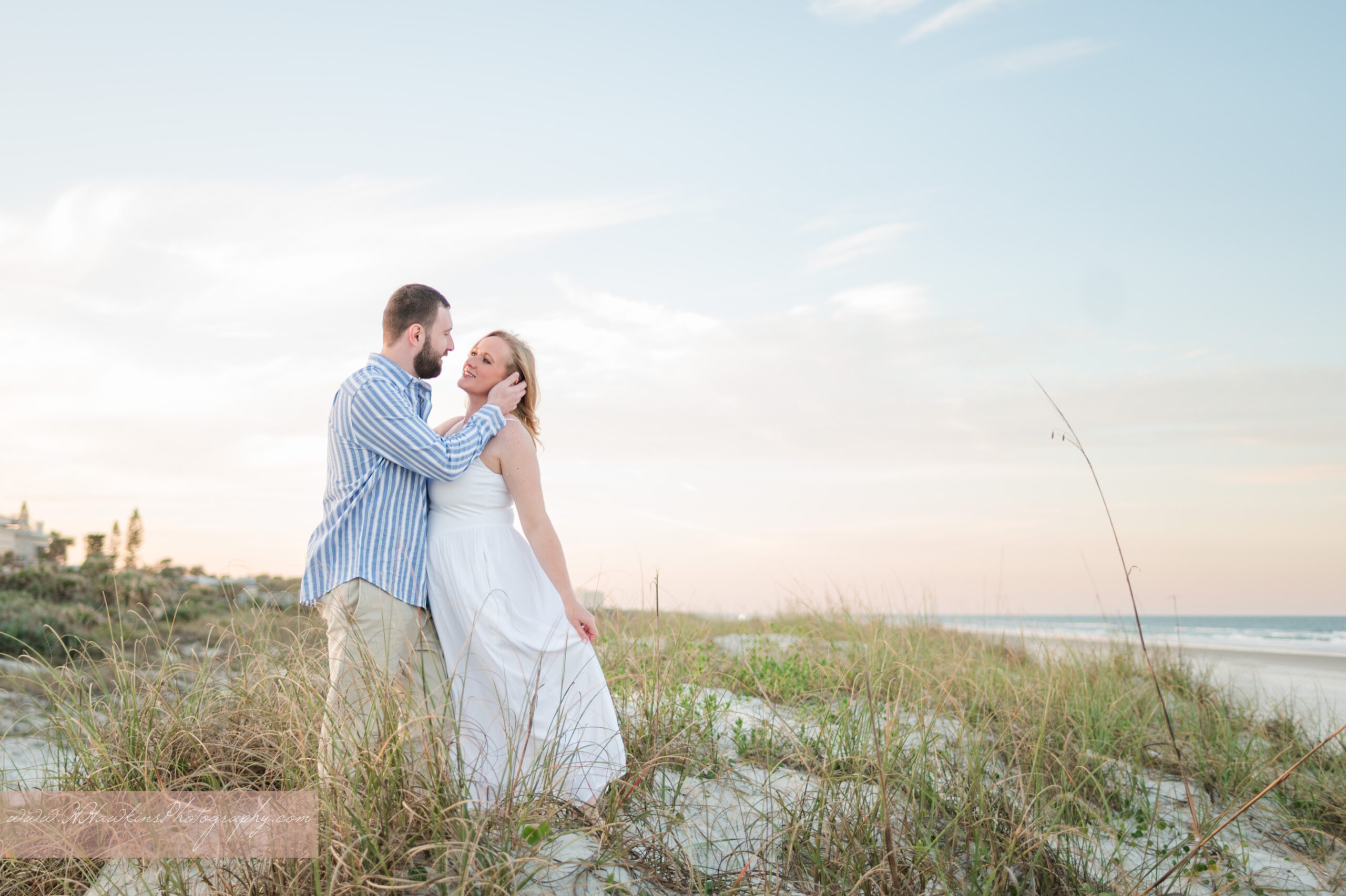 Engaged couple stands on the sand dunes in New Smyrna Beach with the ocean and cotton candy sunset behind them for their their florida beach photos